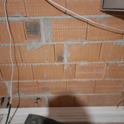 Bogdan Topolanek - Surowa ściana z czerwonej cegły dziurawki w trakcie prac budowlanych, widoczne przewody elektryczne prowadzone po ścianie i puszka instalacyjna, na podłodze białe listwy przypodłogowe, rury...