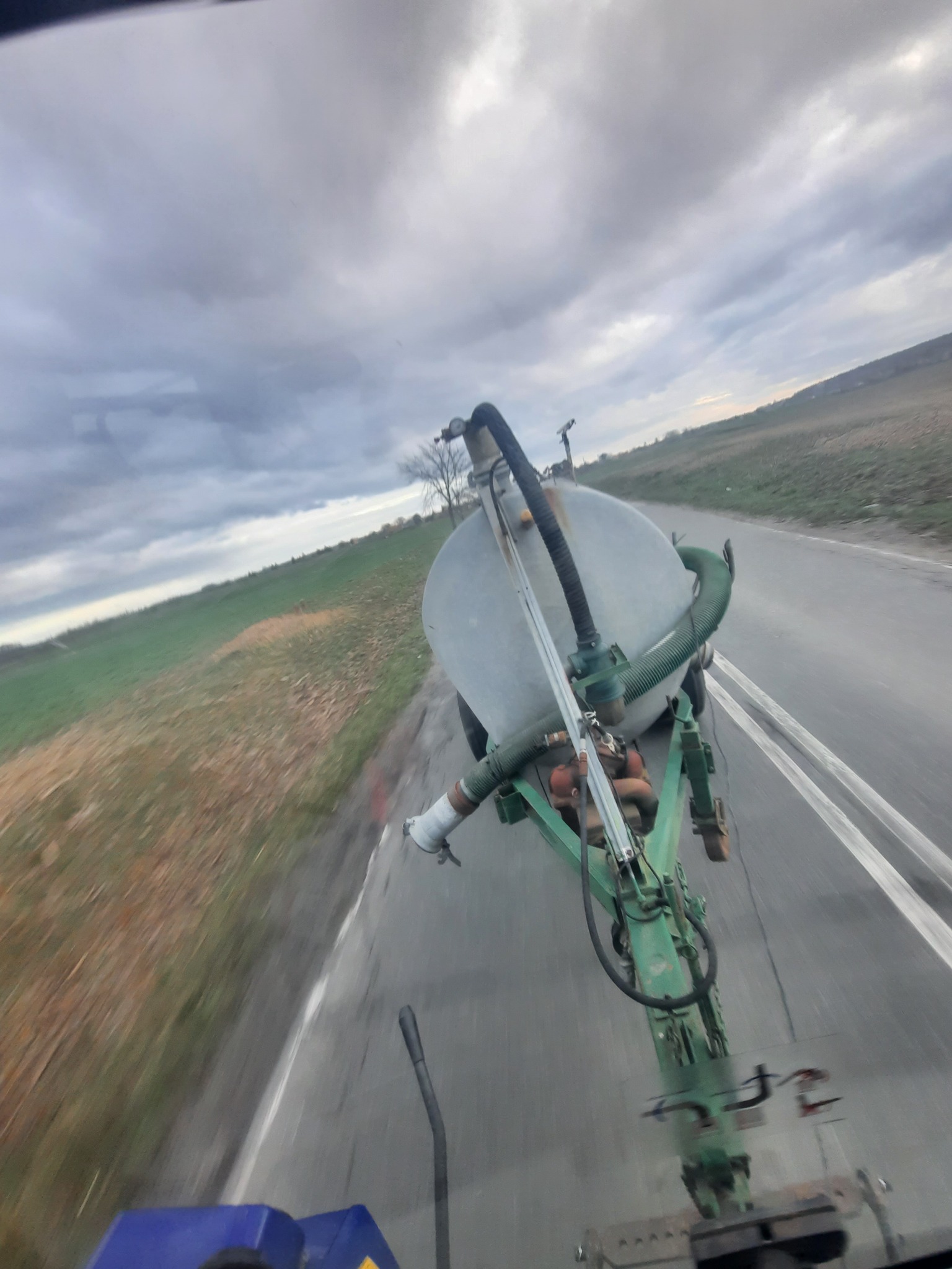 Widok z kabiny ciągnika na przyczepę z zielonym stelażem i szarym zbiornikiem podczas jazdy po asfaltowej drodze, z polami i pochmurnym niebem w tle.