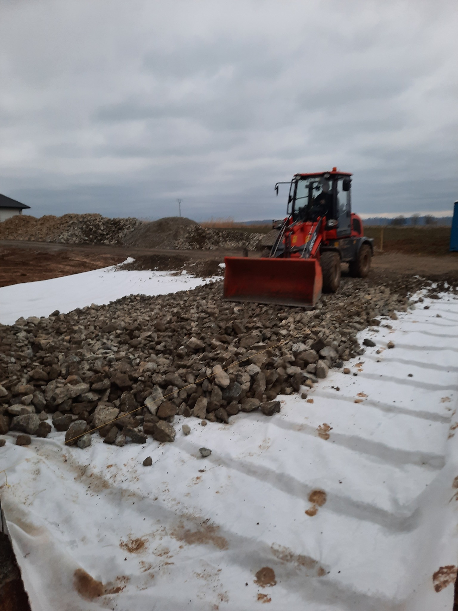 Czerwona ładowarka czołowa rozładowuje kamienie na białą geowłókninę, widoczny operator w kabinie, w tle hałda ziemi i fragment budynku.