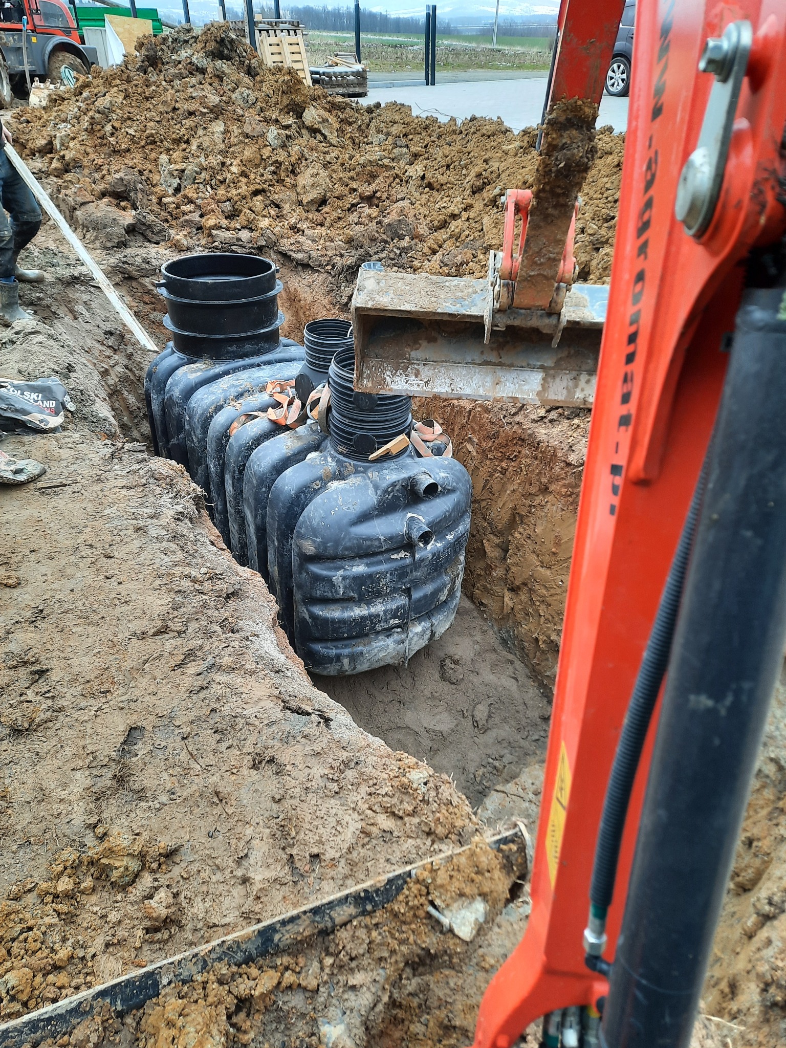 Instalacja zbiornika bezodpływowego w wykopie; widoczny pomarańczowy wysięgnik koparki i sterta ziemi w tle.