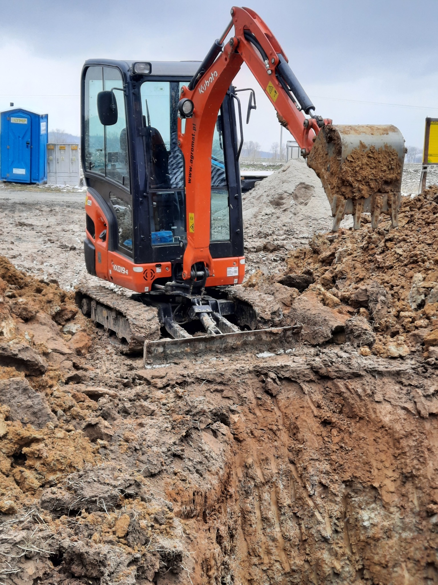 Pomarańczowa mini koparka Kubota z łyżką pełną ziemi, pracująca przy wykopie w ziemi, z widoczną niebieską toaletą Toi Toi w tle.
