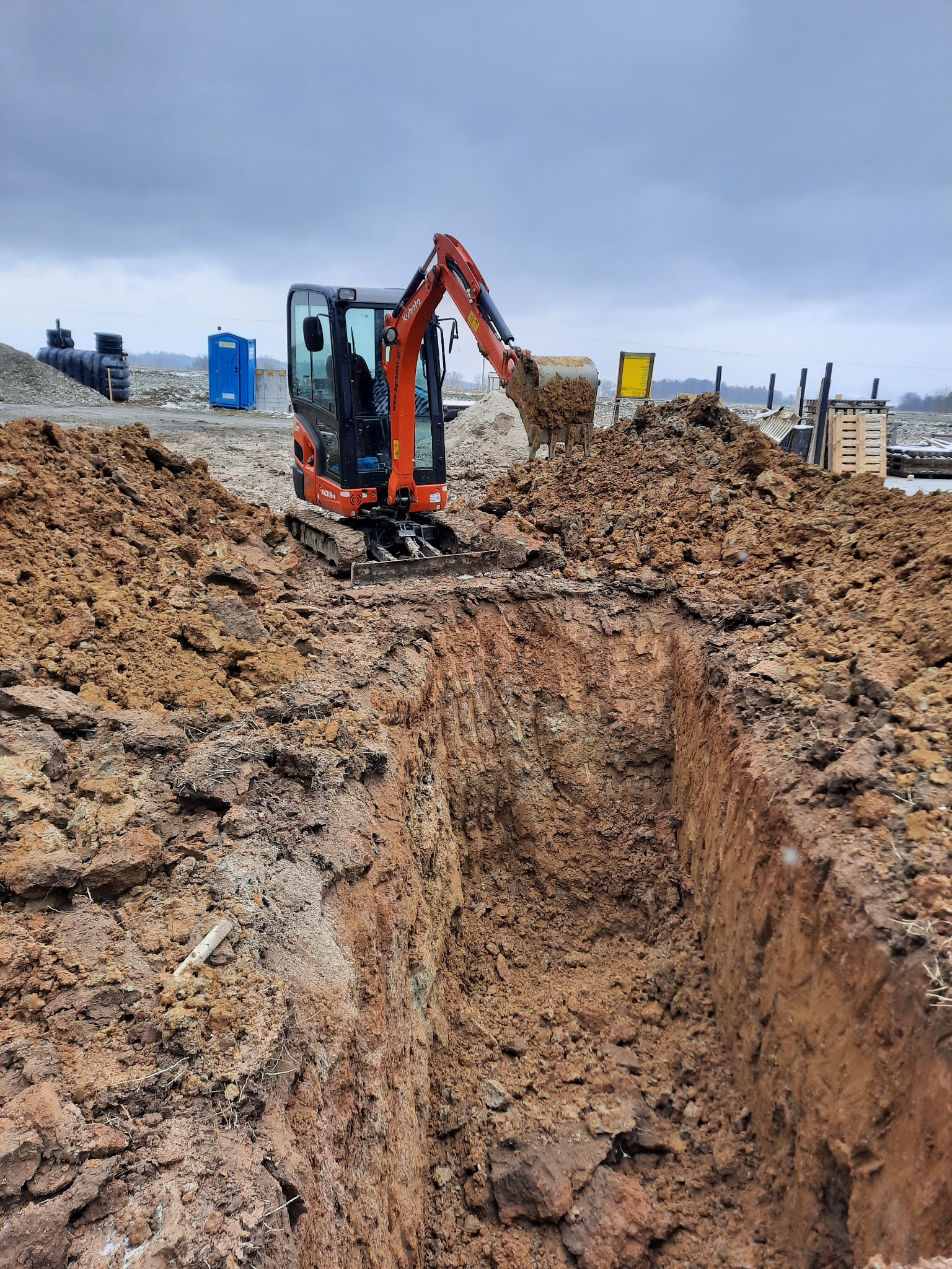 Mała pomarańczowa koparka Kubota na placu budowy, pogłębiająca wąski wykop w gliniastej ziemi, z widocznymi czarnymi rurami i niebieską toaletą TOI-TOI w tle pod pochmurnym niebem.
