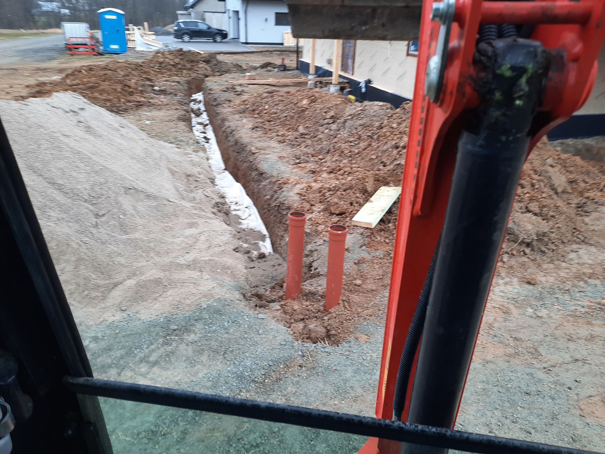 Widok z kabiny koparki na wykopany rów z ułożoną białą geowłókniną i wystającymi rurami kanalizacyjnymi, widoczny plac budowy z przenośną toaletą i budynkiem w tle.