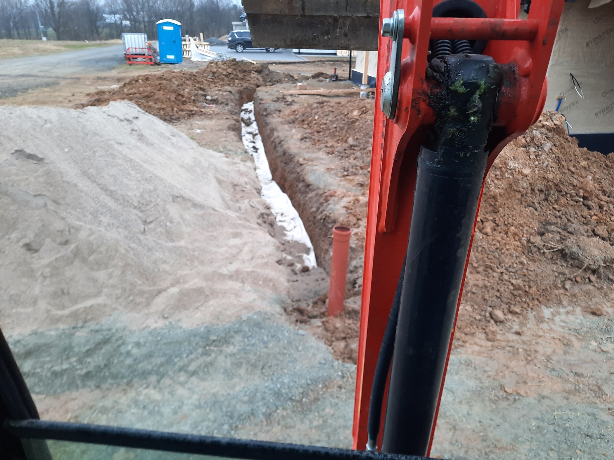 Wykopany wąski rów w ziemi, widok z kabiny koparki na plac budowy z przenośną toaletą w tle.