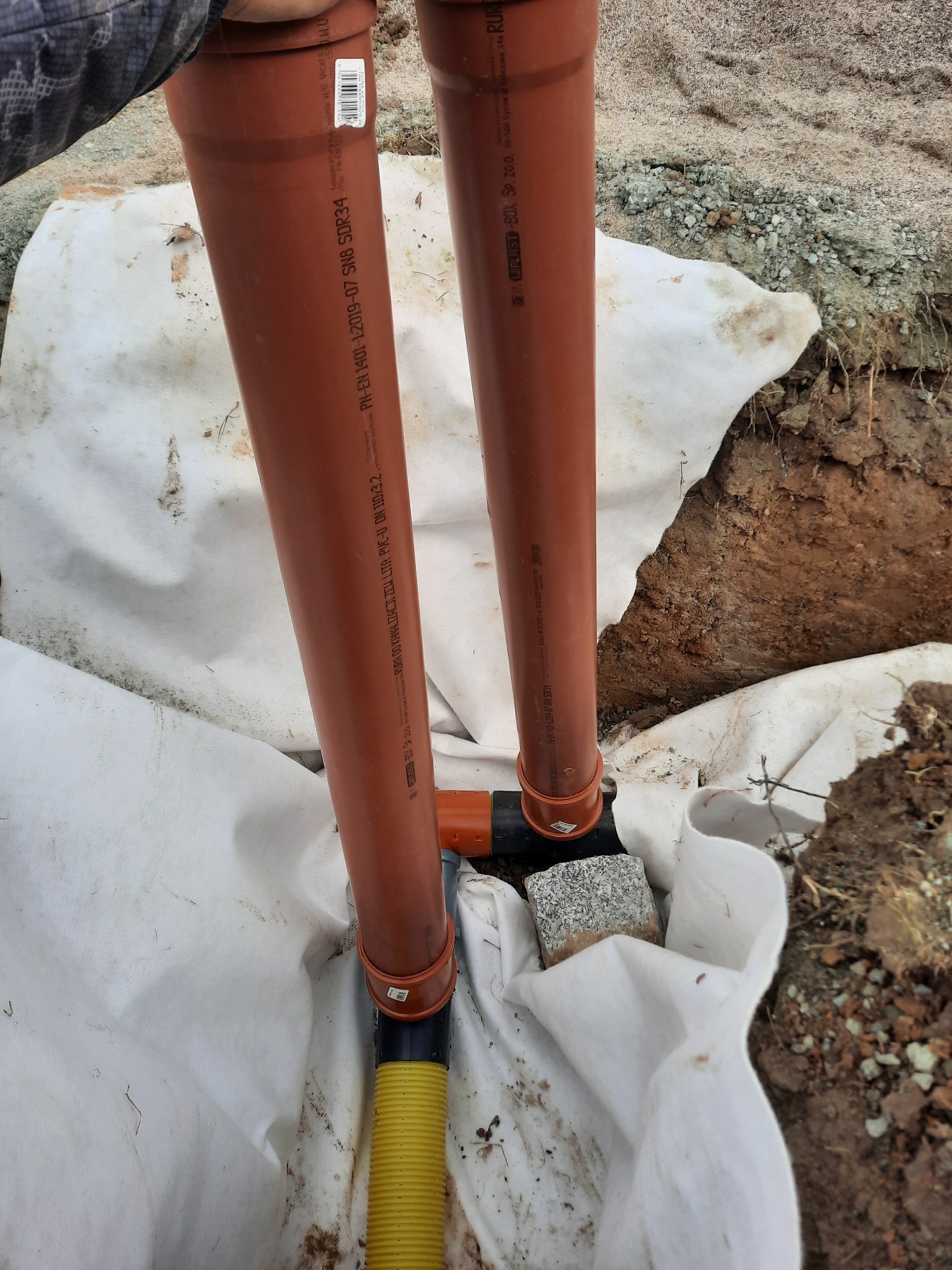 Dwa pionowe brązowe rury kanalizacyjne z tworzywa sztucznego, połączone kształtką, w wykopie wyłożonym białą geowłókniną. Widoczny fragment żółtej rury karbowanej i kamień.  Rury posiadają...