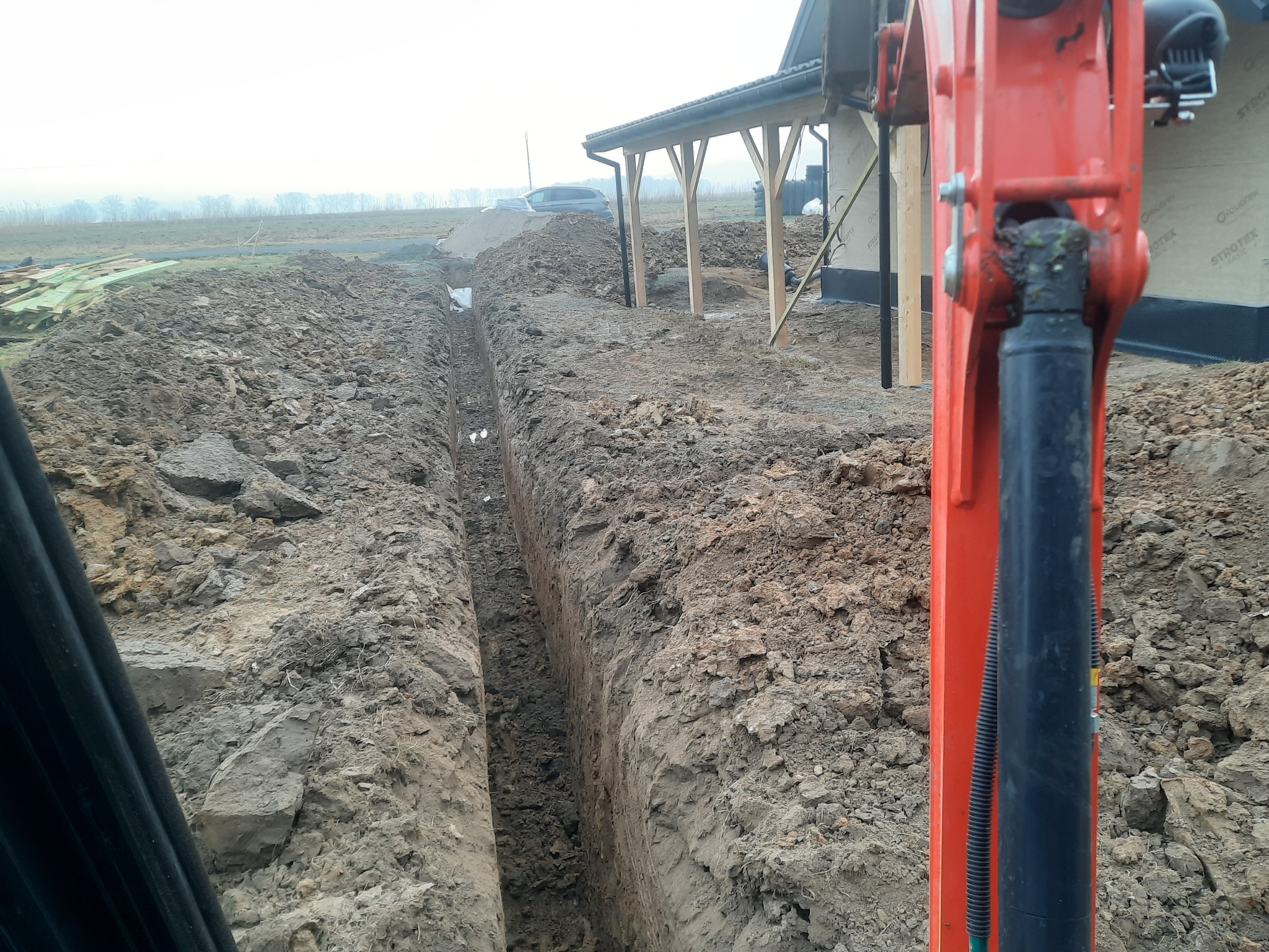 Wykopany wąski rów w ziemi, widoczny z perspektywy operatora koparki, z fragmentem ramienia maszyny na pierwszym planie; w tle zarys domu w trakcie budowy i elementy konstrukcyjne z drewna.