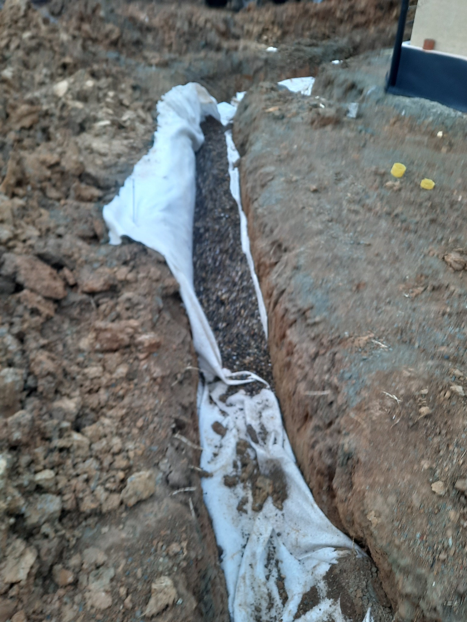 Wykop w ziemi wyłożony geowłókniną wypełniony żwirem, prawdopodobnie drenaż.