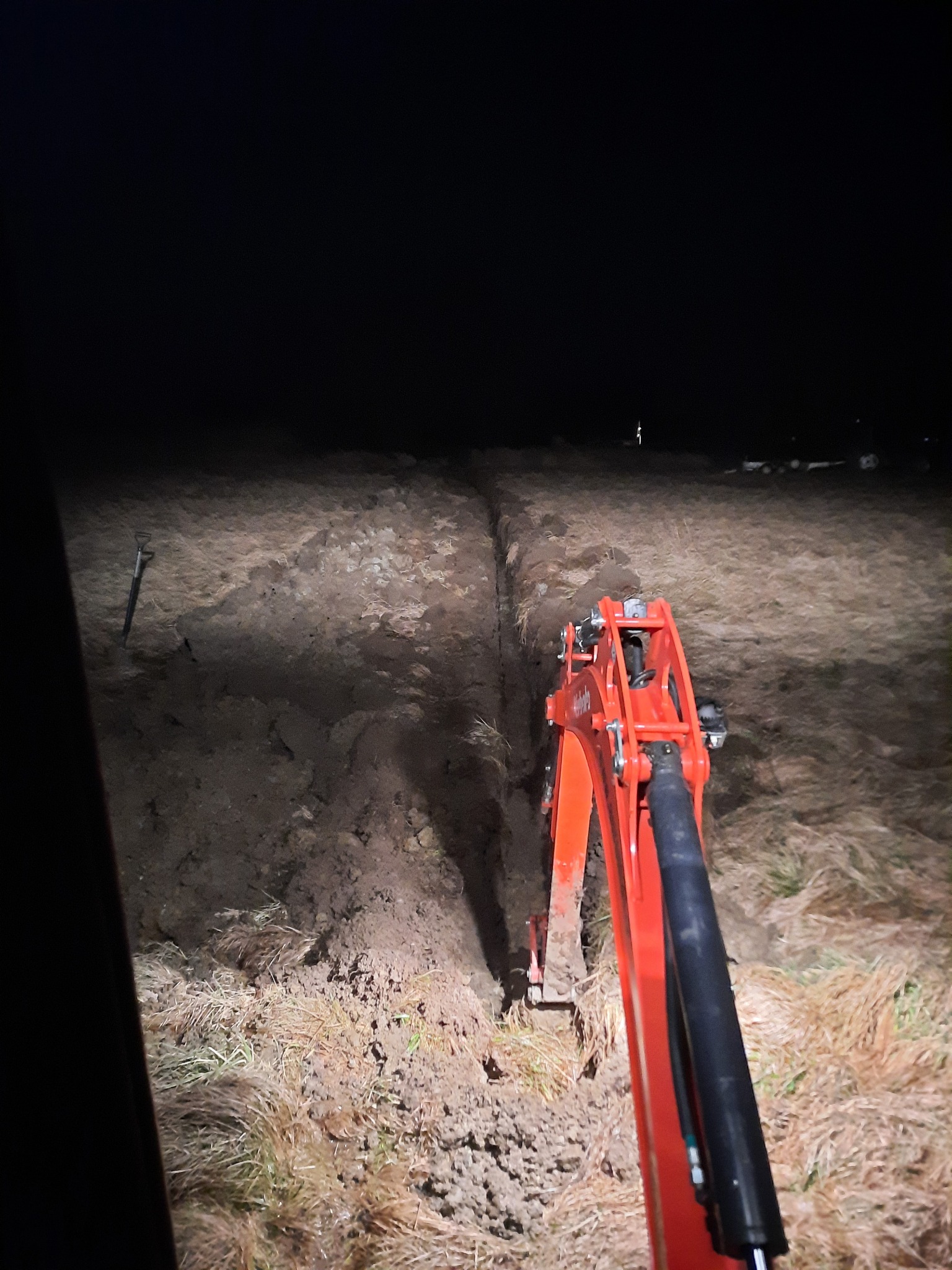Pomarańczowa koparka Kubota w trakcie wykonywania wykopu pod instalację w słabym oświetleniu nocnym, widoczna długa bruzda w ziemi i trawa na brzegach.