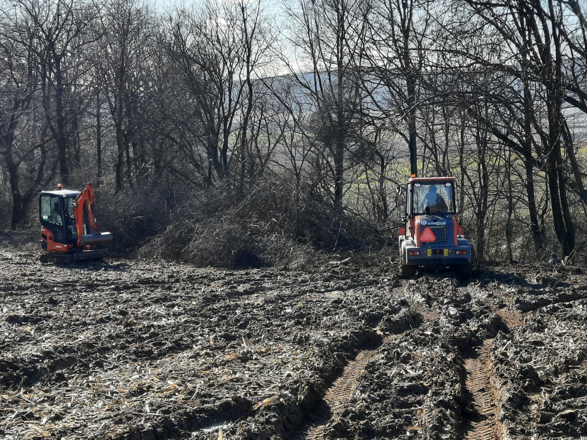 Dwa traktory na polu po wycince drzew, widoczne ślady opon w błotnistym terenie, w tle drzewa bez liści i zarys gór.