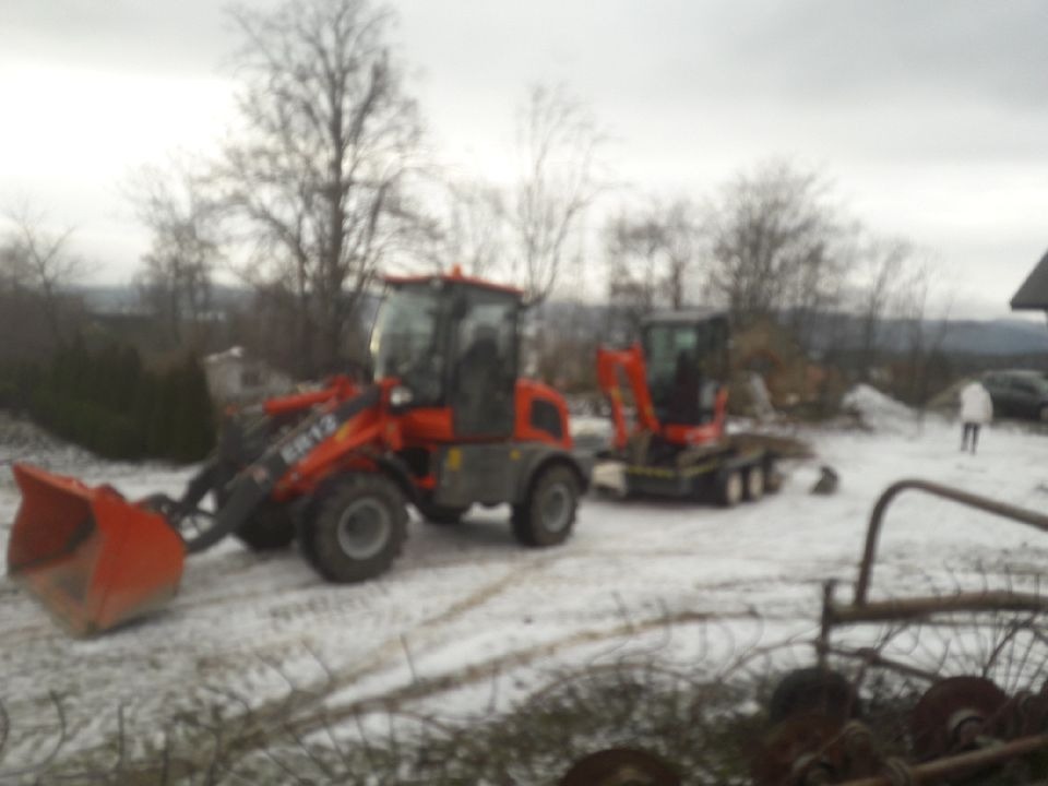 Pomarańczowa ładowarka ER12 i mała koparka na śniegu, widoczne drzewa w tle i osoba w białej kurtce.