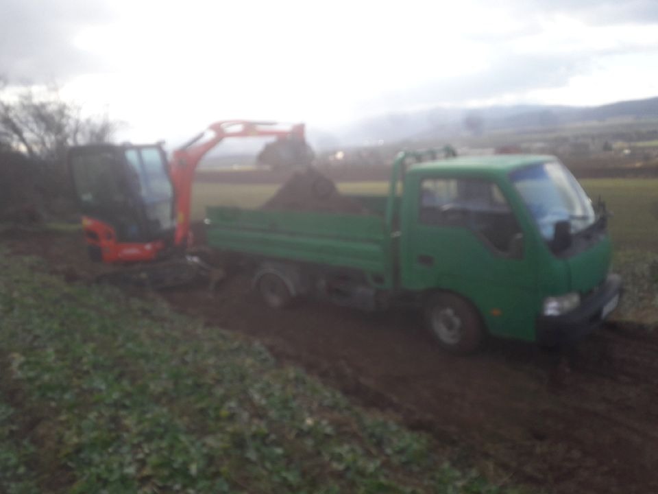 Mała koparka ładuje ziemię do zielonej ciężarówki na rozmokłym terenie, w tle widoczne wzgórza i pochmurne niebo.