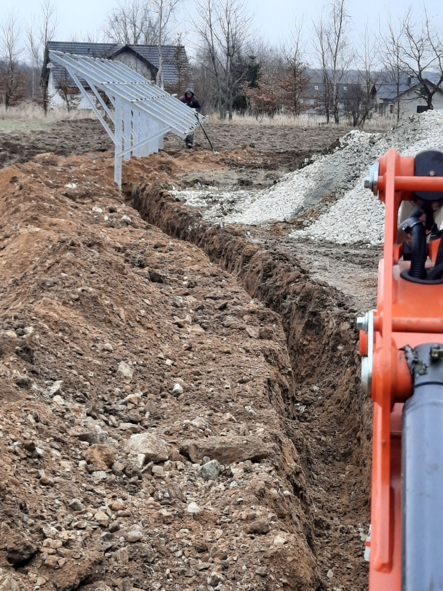 Wąski wykop w ziemi, widoczny szkielet konstrukcji wsporczej pod panele słoneczne, pracownik z łopatą w tle i fragment pomarańczowej maszyny budowlanej po prawej stronie.