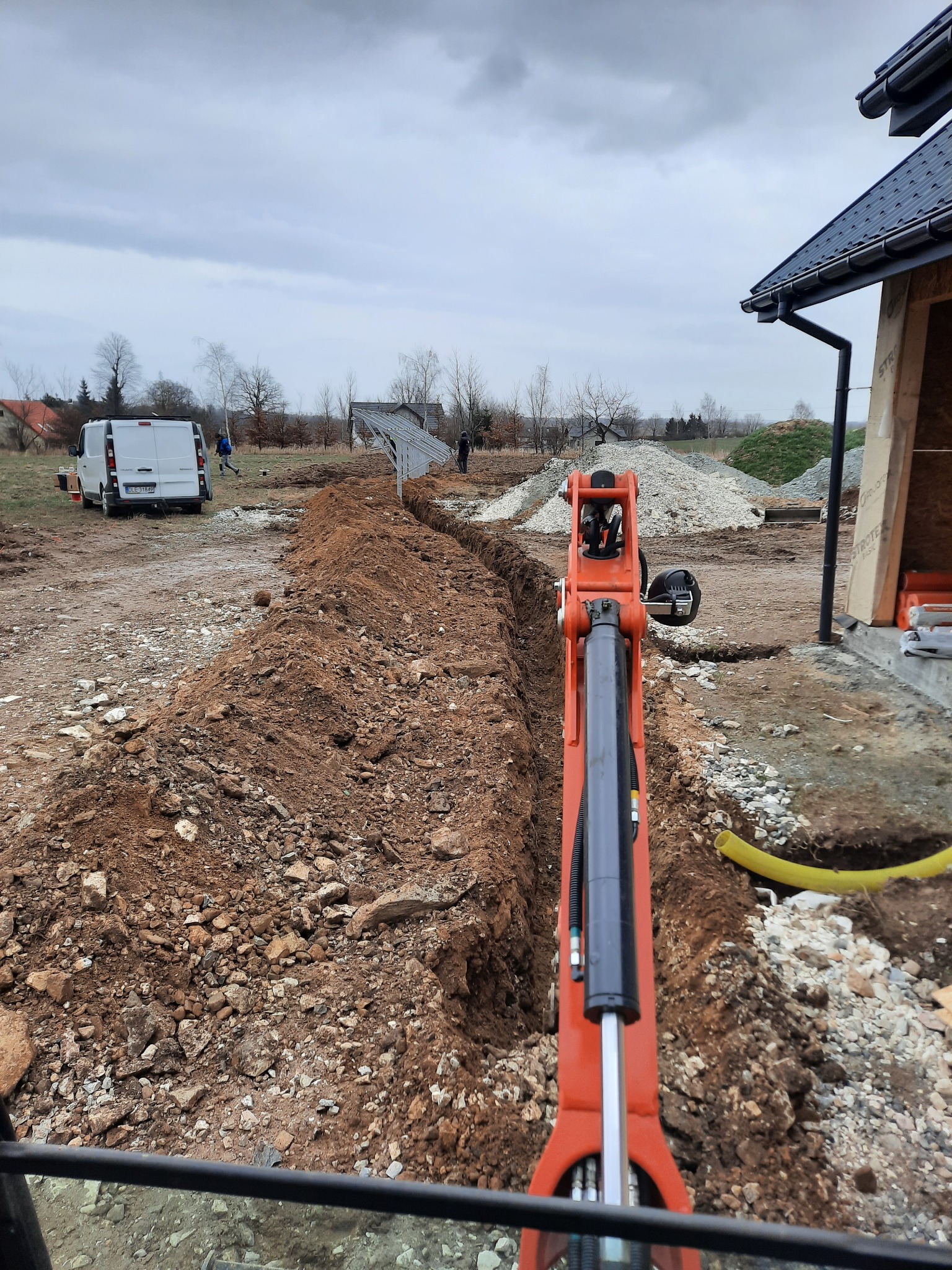 Wykopany wąski rów w Bielawie, widziany z perspektywy operatora pomarańczowej mini koparki. W tle biały bus i częściowo zmontowana konstrukcja, prawdopodobnie pod panele fotowoltaiczne.