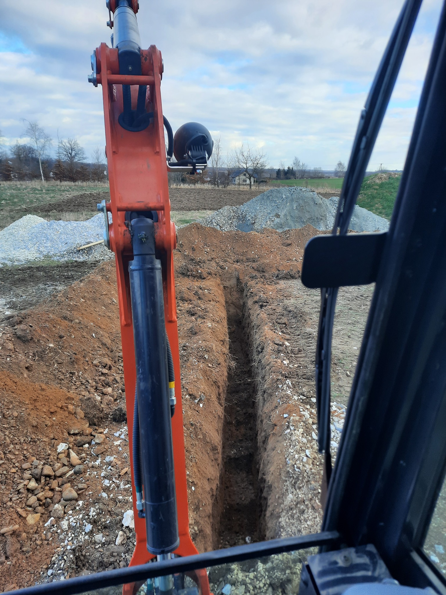 Wąski wykop w ziemi widziany z kabiny pomarańczowej koparki, w tle hałdy żwiru i pochmurne niebo.