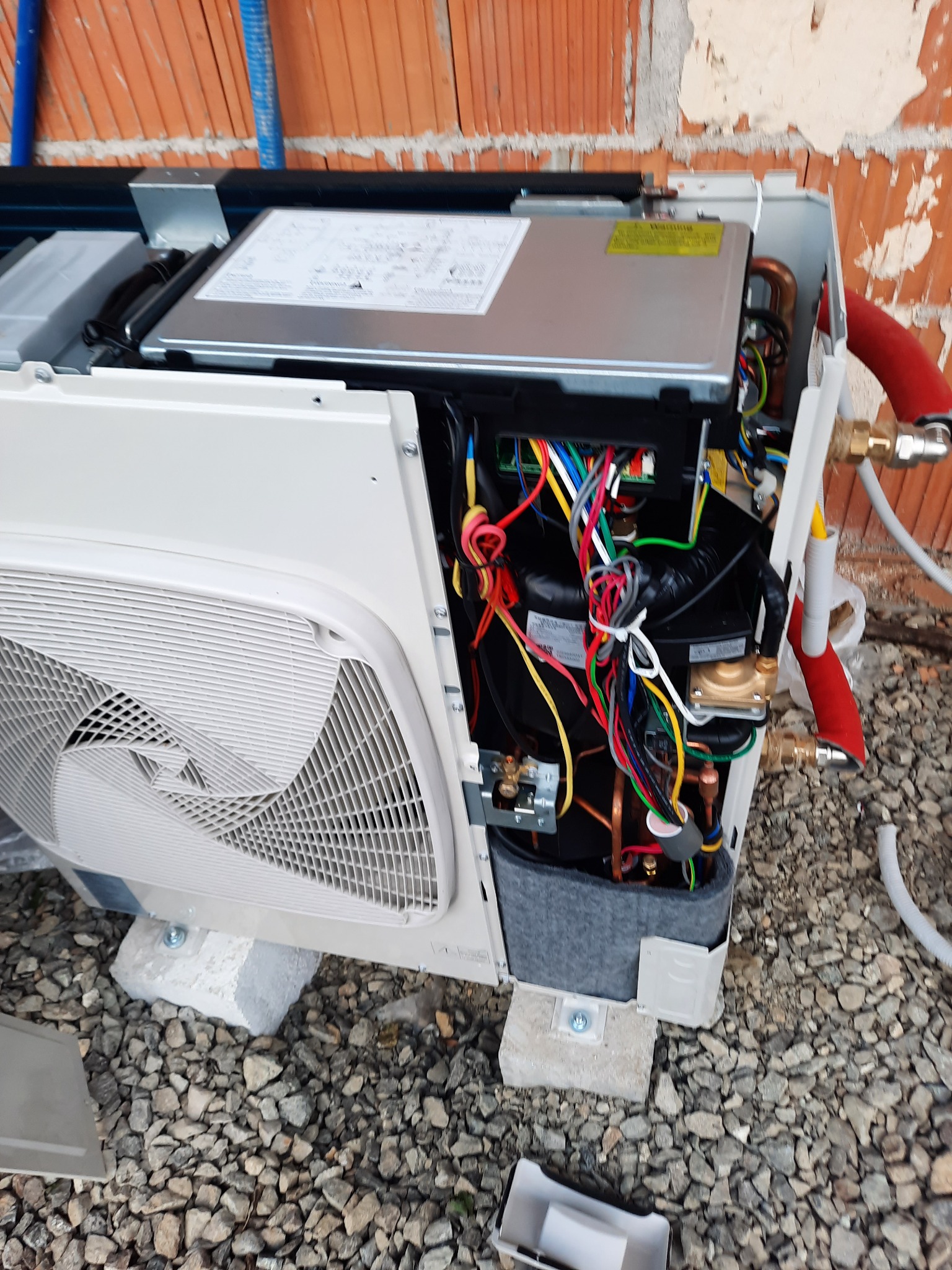 Otwarta obudowa zewnętrznej jednostki pompy ciepła, widoczne wewnętrzne komponenty, przewody elektryczne, rury miedziane, zamocowana na betonowych bloczkach na żwirowym podłożu, z fragmentem ściany...