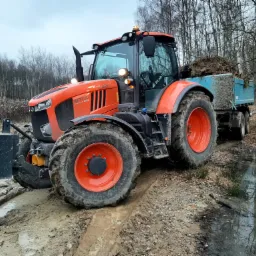 Pomarańczowy ciągnik Kubota M7173 z przyczepą wypełnioną gałęziami, stoi na błotnistej drodze w lesie, odbicie drzew w wodzie.