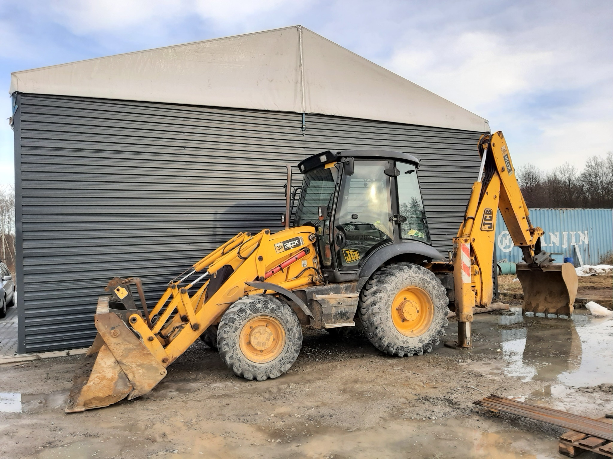 Żółta koparko-ładowarka JCB 3CX zaparkowana na błotnistym terenie przed szarą halą z białą plandeką, widoczne ślady pracy i mokra nawierzchnia.
