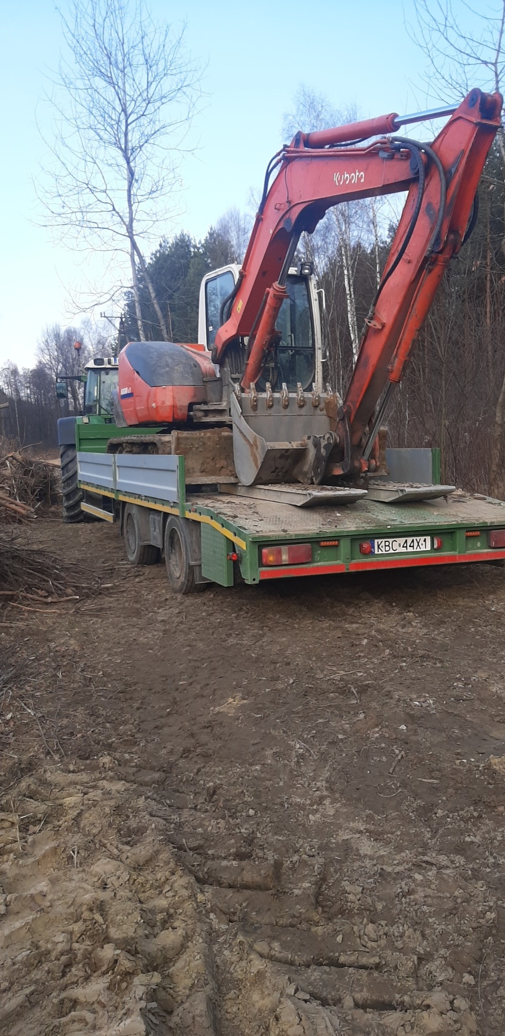 Pomarańczowa koparka Kubota na zielonej lawecie transportowana traktorem w Rzezawie, widoczne ślady opon na błotnistym terenie.