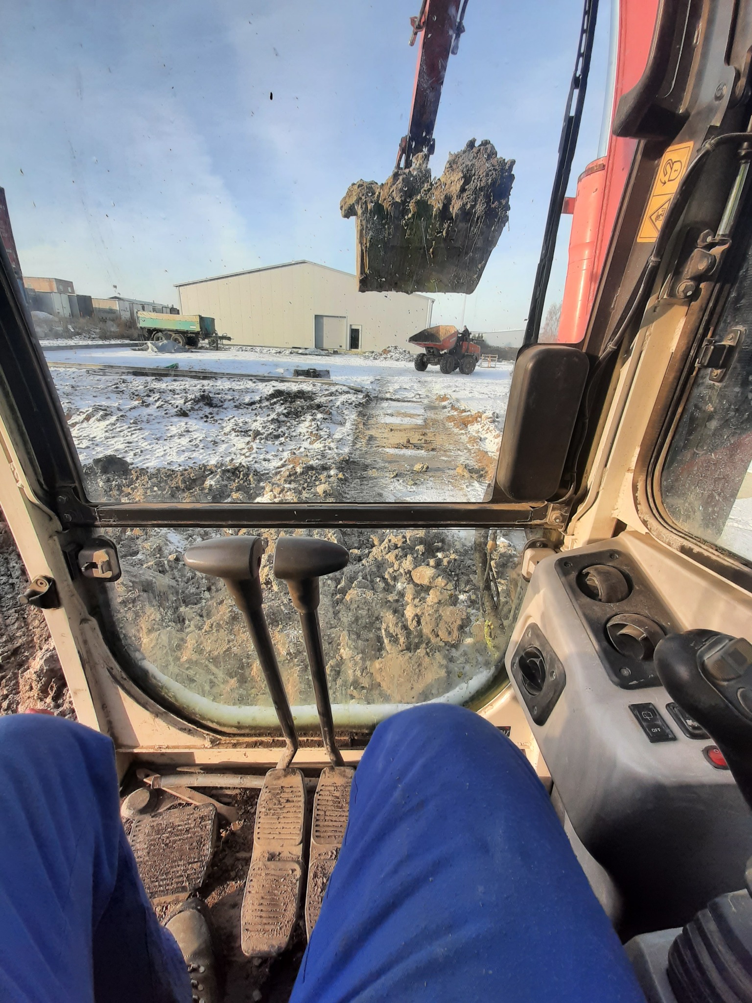 Widok z kabiny koparki podczas pracy w zimowych warunkach, łyżka nabiera ziemię, w tle budynek i wywrotka.