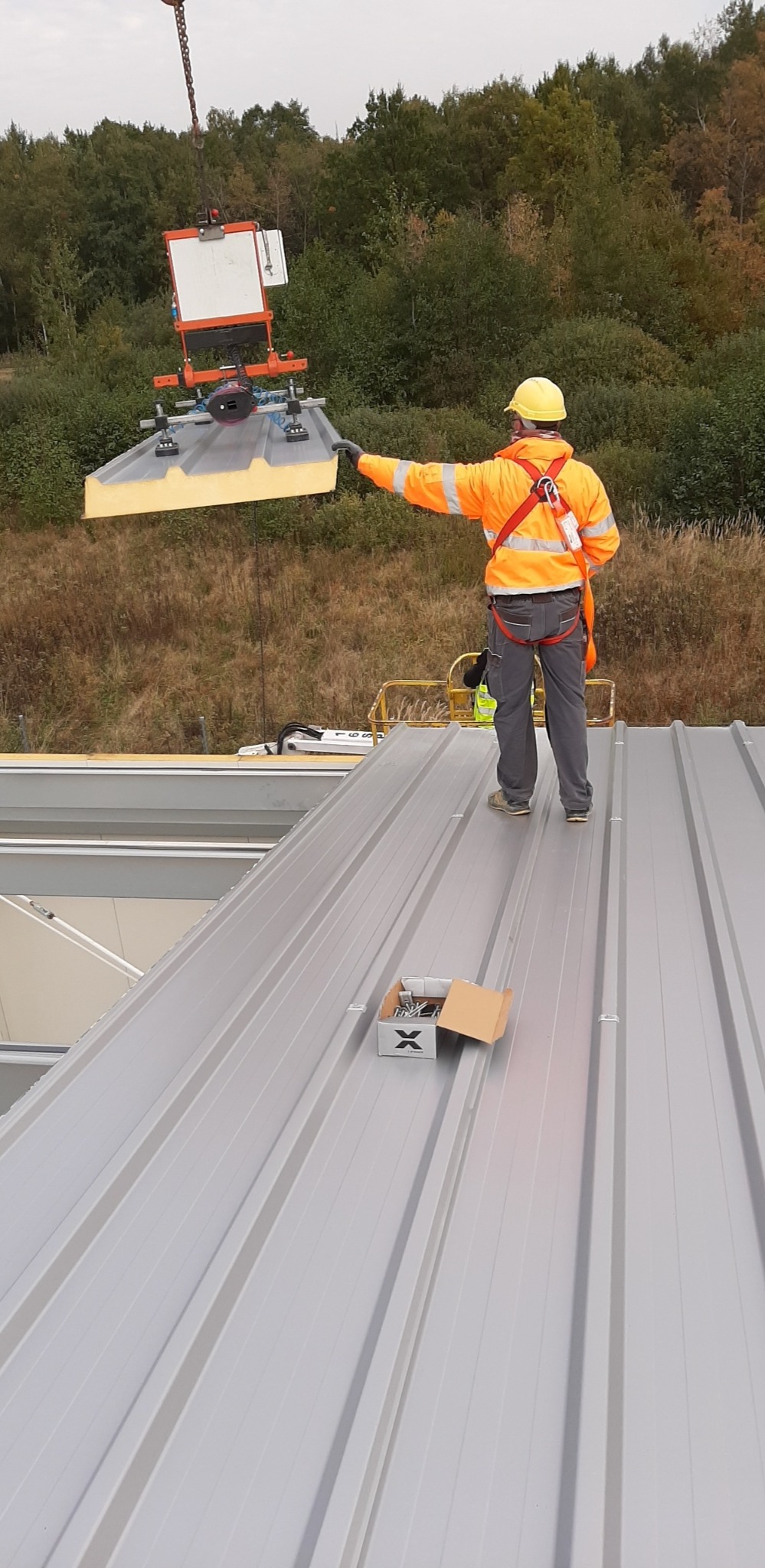Pracownik w pomarańczowej kurtce i żółtym kasku, zabezpieczony uprzężą, montuje metalowe panele dachowe przy użyciu przyssawki podwieszonej do dźwigu. Na dachu widoczny karton z wkrętami.