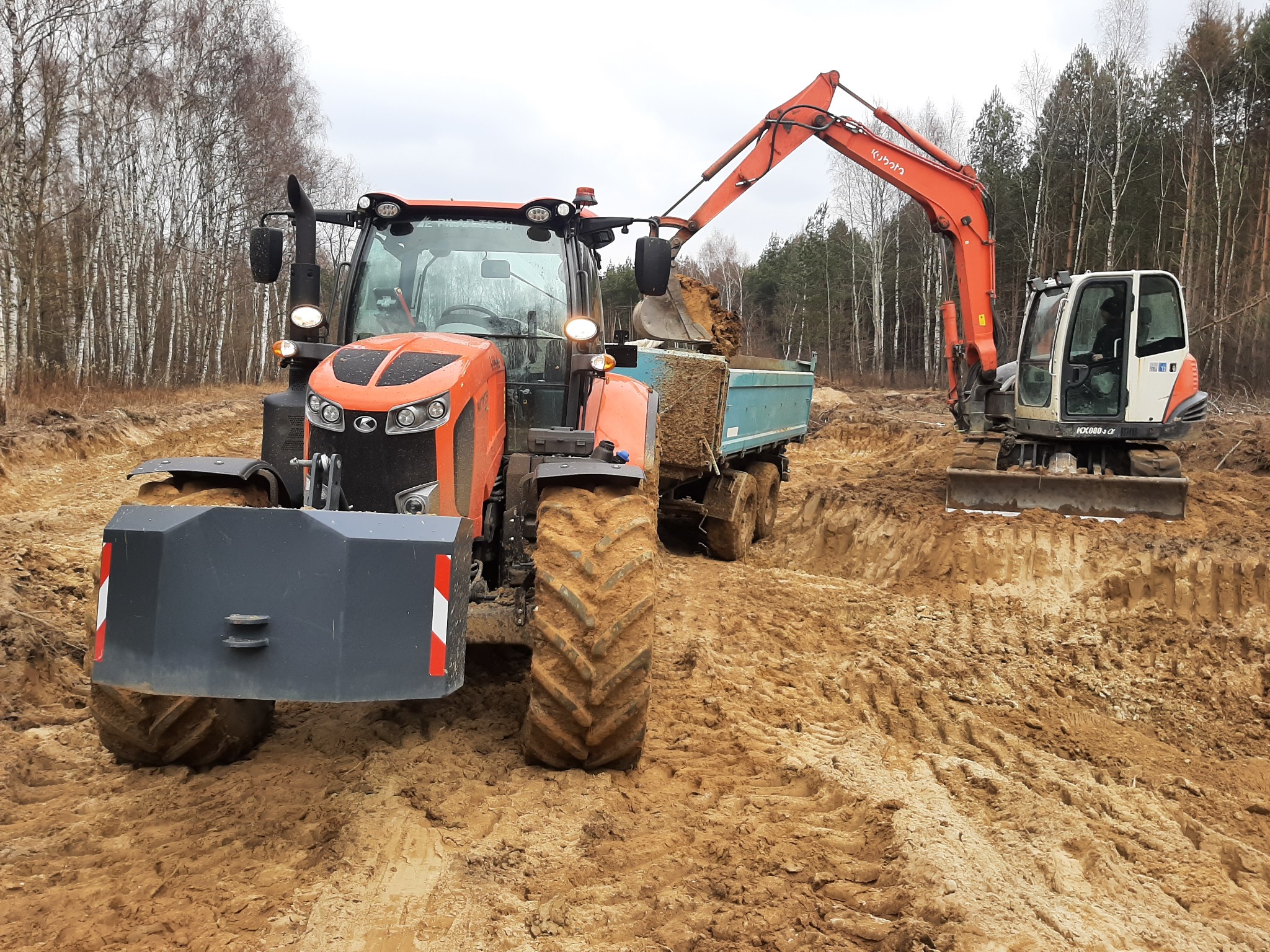 Pomarańczowy traktor i minikoparka Kubota podczas prac ziemnych na placu budowy z drzewami w tle. Koparka nabiera ziemię do przyczepy traktora.