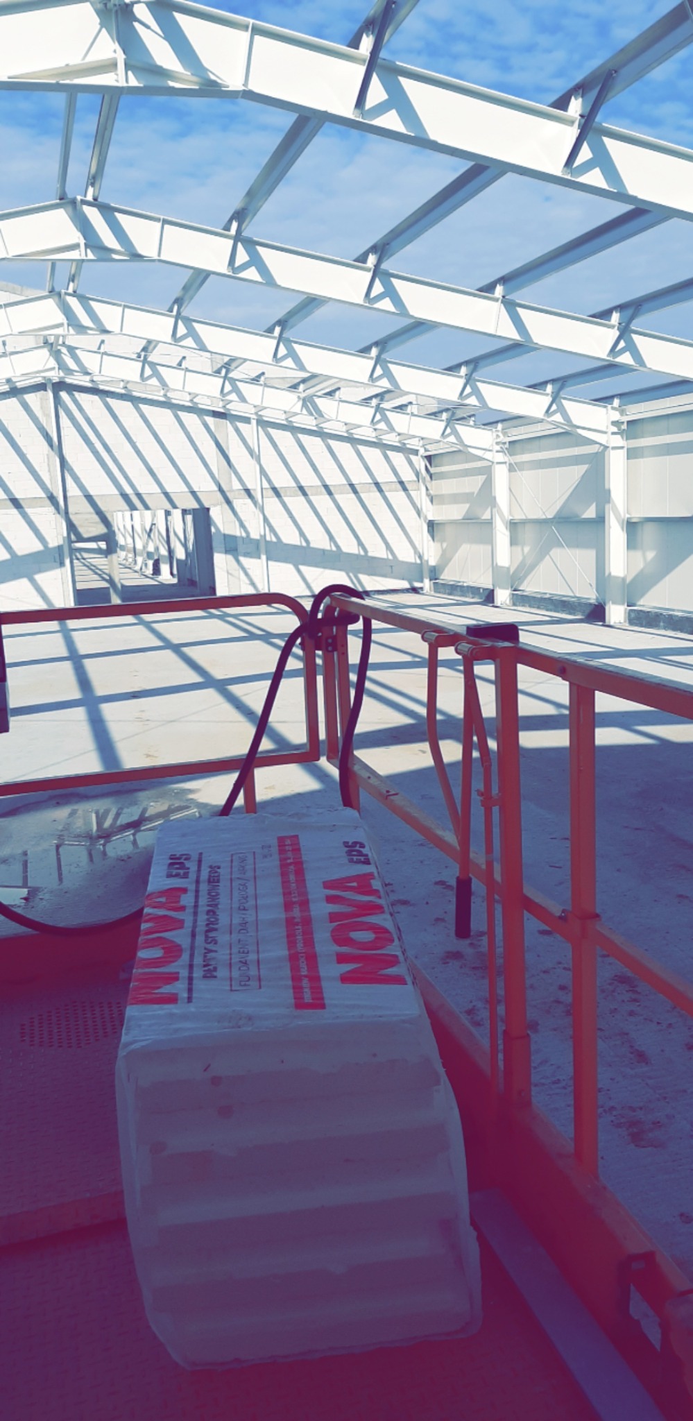 Konstrukcja stalowa hali w trakcie budowy, widoczne białe belki stropowe i ściany, pomarańczowe barierki oraz paczka styropianu NOVA EPS na podłodze.