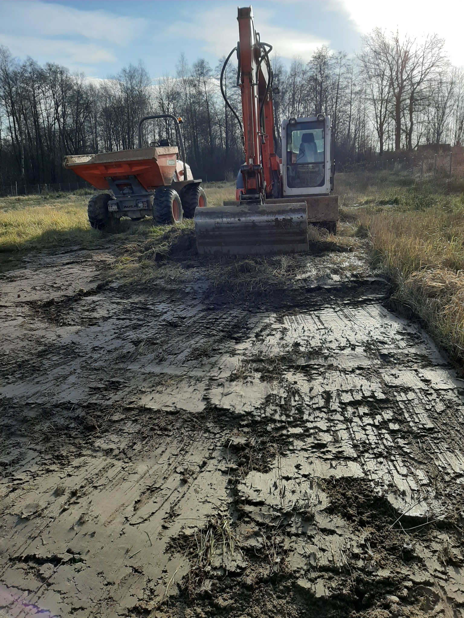Pomarańczowa wywrotka i koparka na placu budowy, świeżo wyrównana ziemia z widocznymi śladami opon i łyżki, w tle drzewa bez liści i błękitne niebo.