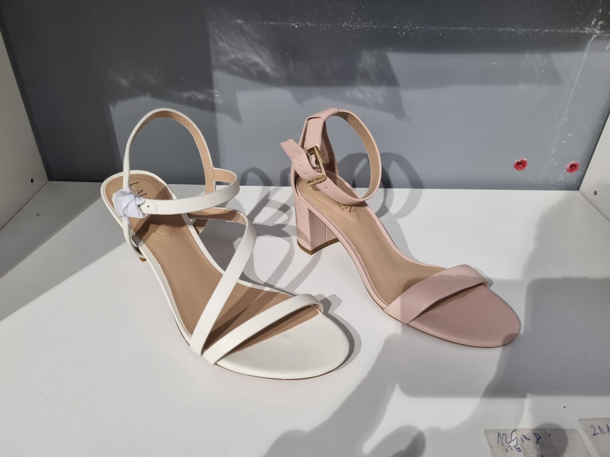 Dwa różne modele damskich sandałów na obcasie, białe i pudrowo różowe, prezentowane na półce w sklepie w Warszawie, z widocznymi cenami na kartkach w tle.