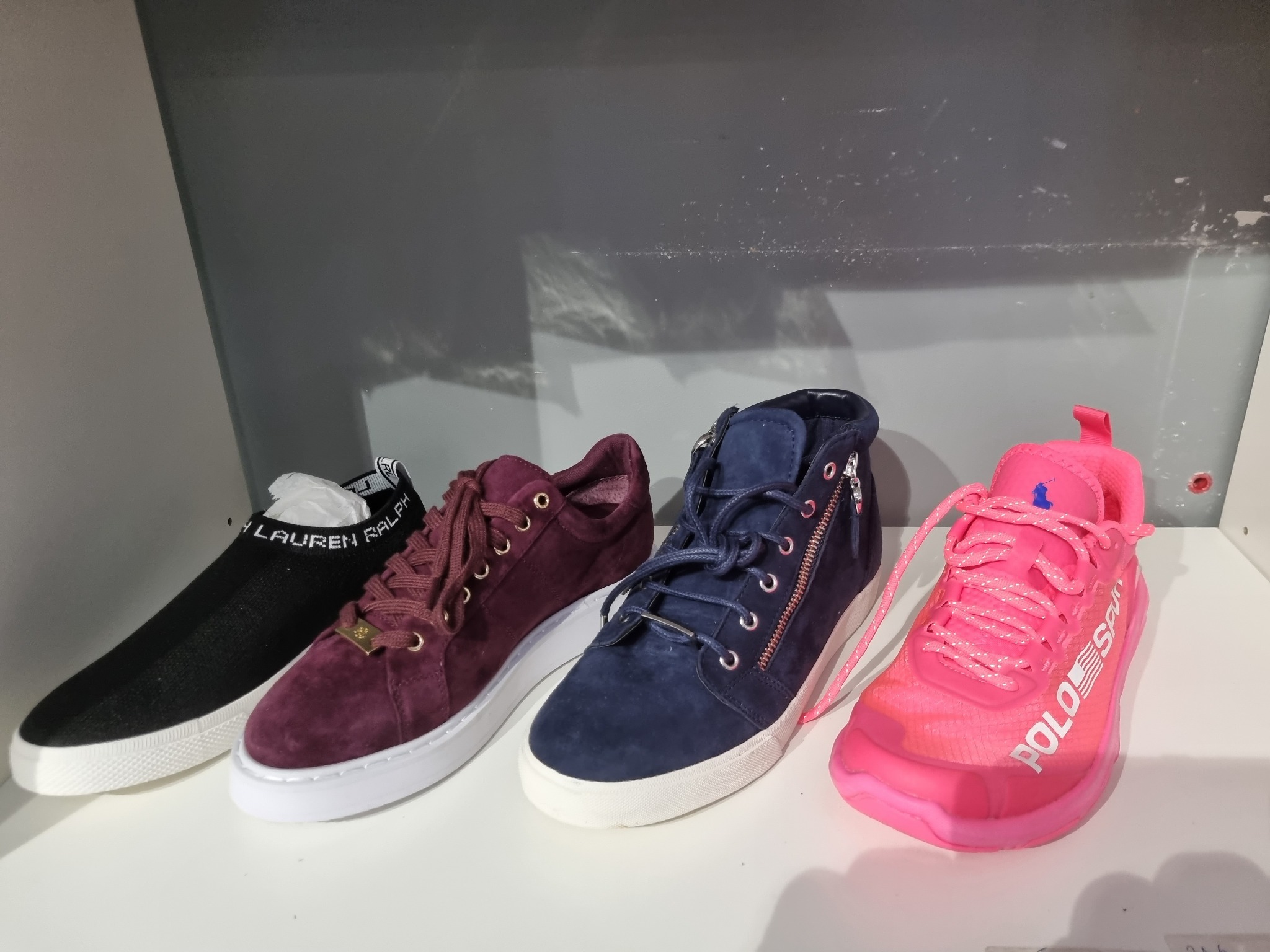 Cztery pary butów sportowych: czarne wsuwane z napisem, bordowe sznurowane zamszowe, granatowe sznurowane z zamkiem i różowe sportowe z napisem Polo Sport, ustawione na białej półce na tle szarej...