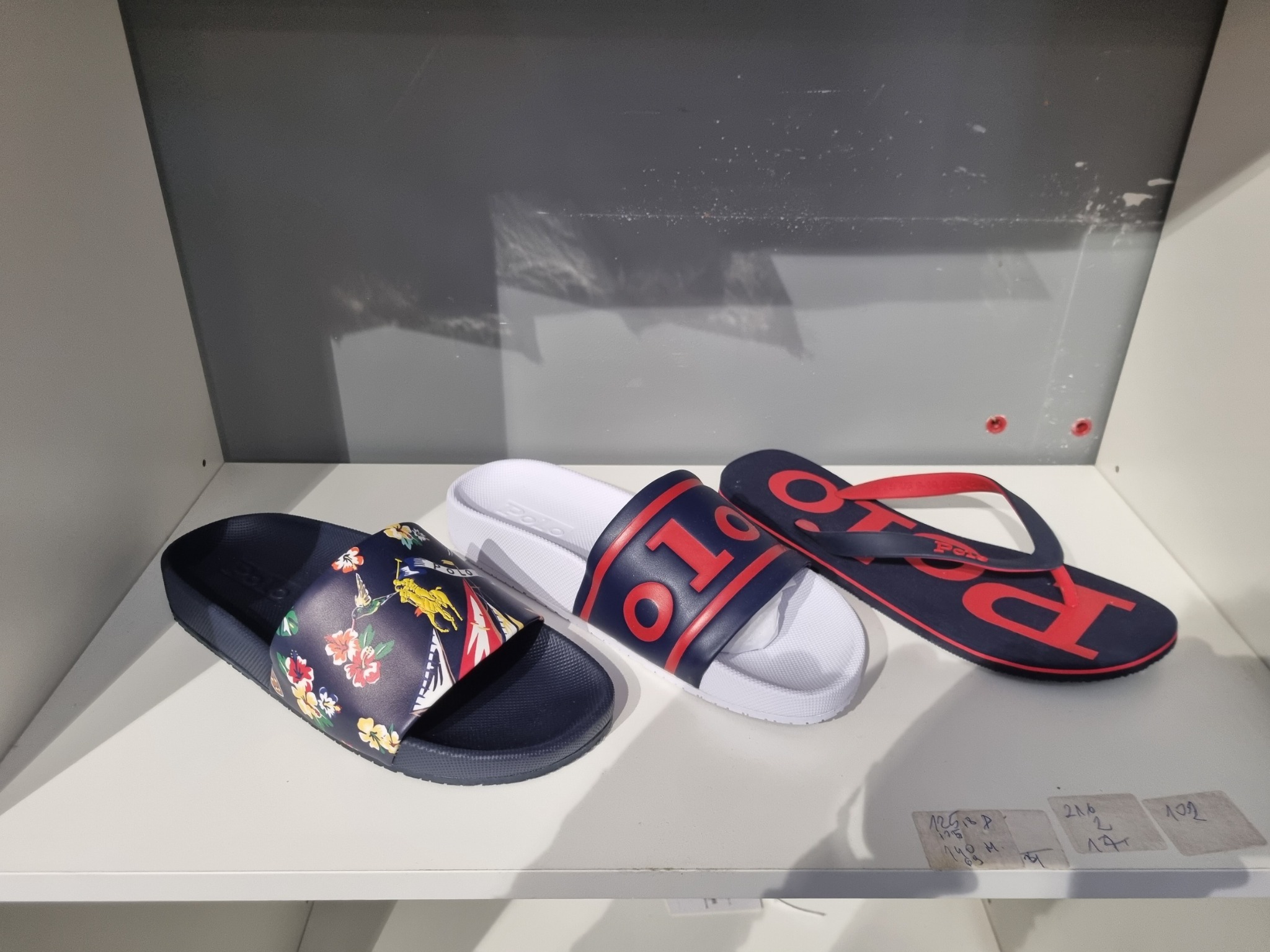 Trzy pary klapek marki Polo Ralph Lauren na białej półce: granatowe z motywem kwiatowym i jeźdźcem, białe z granatowym logo oraz granatowo-czerwone japonki z logo.