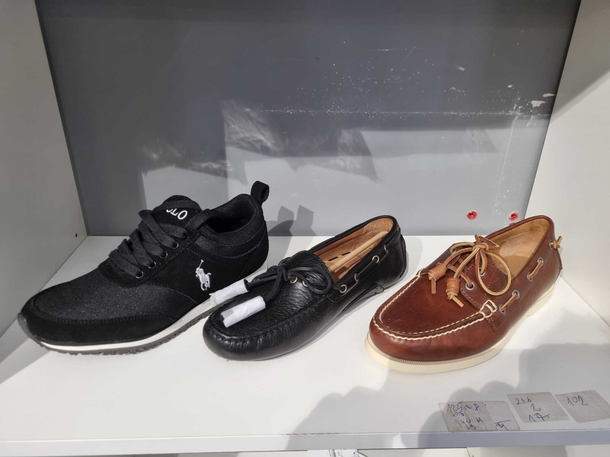 Trzy pary butów męskich: czarne sportowe z logo, czarne skórzane mokasyny z frędzlami i brązowe skórzane sznurowane buty żeglarskie, ustawione na białej półce na tle szarej ściany.