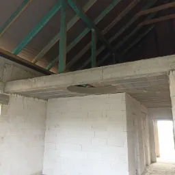 Wnętrze surowego domu w trakcie budowy: białe ściany z betonu komórkowego, strop z prefabrykatów, drewniana konstrukcja dachu z membraną paroprzepuszczalną, widoczne przewody elektryczne na podłodze.