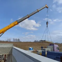 Żółty dźwig podnosi betonowy strop kanałowy na budowie domu z bloczków YTONG, widoczny przenośny toi-toi i ciężarówka