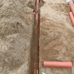 Wykop w piasku z ułożonymi rurami kanalizacyjnymi z tworzywa sztucznego, widoczny poziomica, szpadel i młotek.