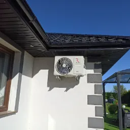 Biała jednostka zewnętrzna klimatyzatora Rotenso zamontowana na elewacji domu z oknem i czarnym dachem w słoneczny dzień.