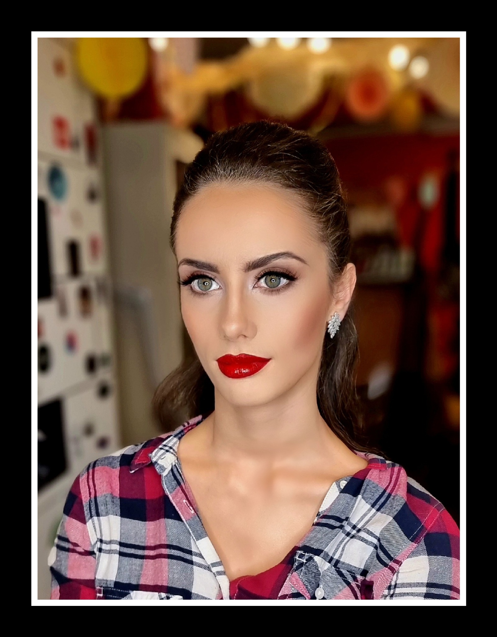 Portret kobiety z profesjonalnym makijażem, w tym intensywnie czerwone usta, podkreślone oczy i delikatny kontur twarzy, uzupełniony błyszczącymi kolczykami i koszulą w kratę.