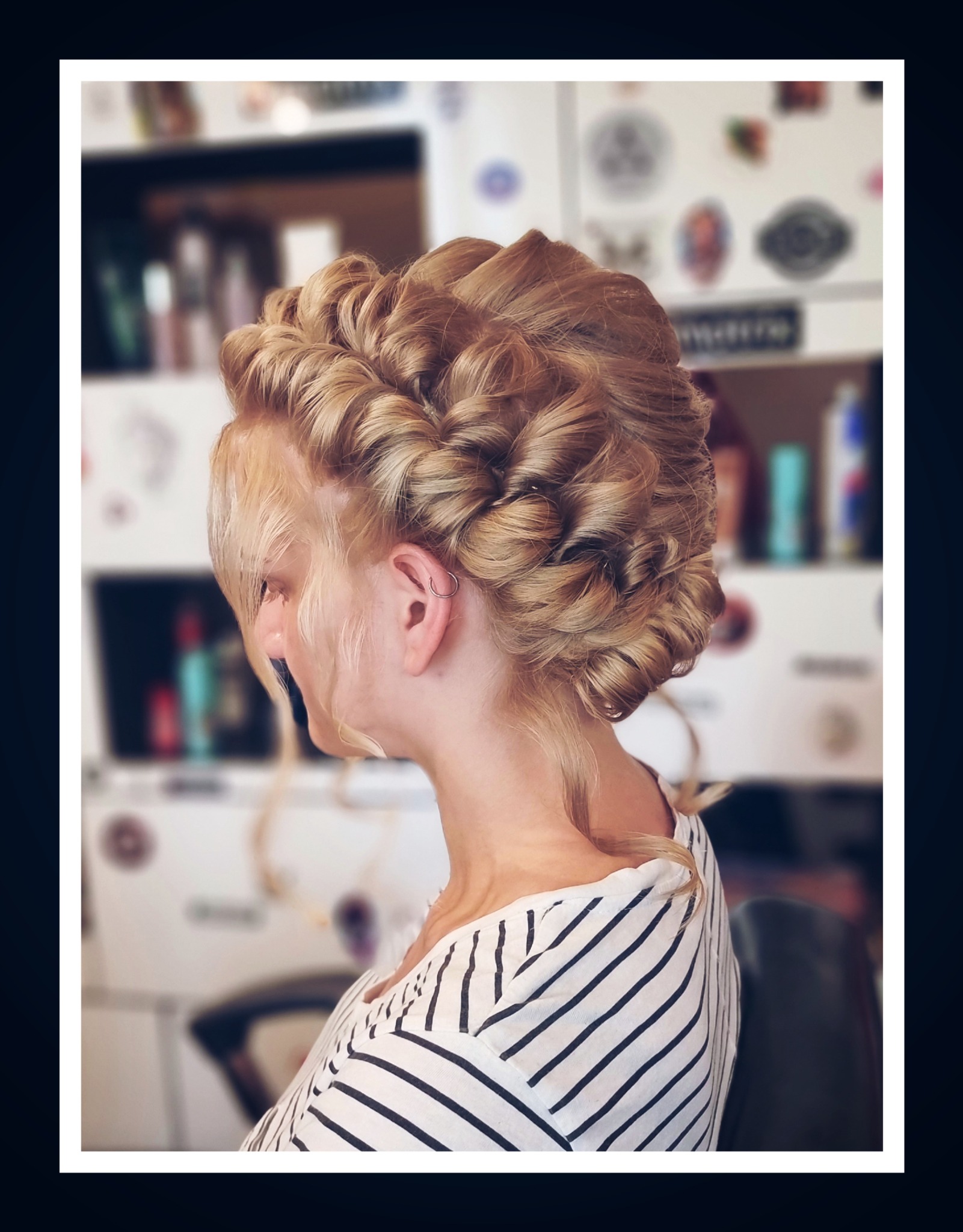 Blond upięcie włosów z warkoczami i lokami, widok z profilu, kolczyk w uchu, bluzka w paski, salon fryzjerski w tle.