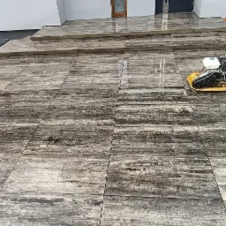 Nowo ułożona, brązowo-szara kostka brukowa przed nowoczesnym domem z szarą stolarką, zagęszczarka wibracyjna na powierzchni, widoczne niedokończone podłączenia elektryczne na elewacji.