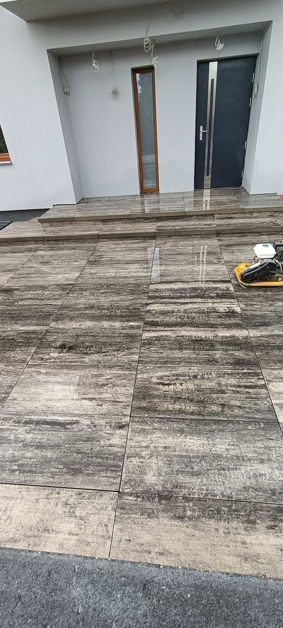 Nowo ułożona, brązowo-szara kostka brukowa przed nowoczesnym domem z szarą stolarką, zagęszczarka wibracyjna na powierzchni, widoczne niedokończone podłączenia elektryczne na elewacji.