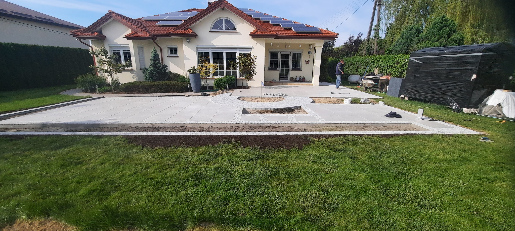 Budowa tarasu z jasnoszarych płyt i kostki granitowej na przygotowanym podłożu, widoczni pracownicy, dom z panelami słonecznymi w tle.