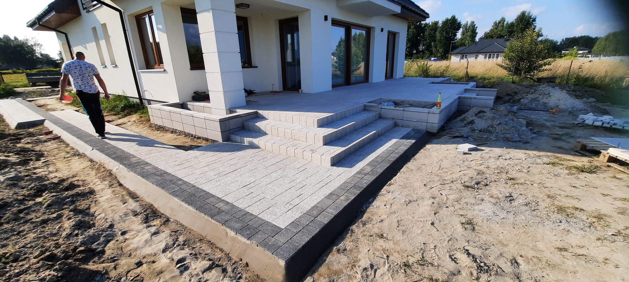 Nowoczesny taras z kostki brukowej w odcieniach szarości, z niskimi schodami prowadzącymi do domu, widoczne niedokończone prace ziemne i palety z kostką w tle.