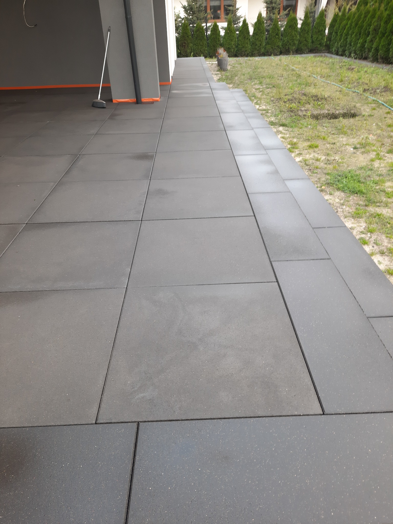 Ułożona, szara kostka brukowa na zewnątrz budynku, przy krawędzi trawnika z tujami, z widoczną szczotką opartą o ścianę.