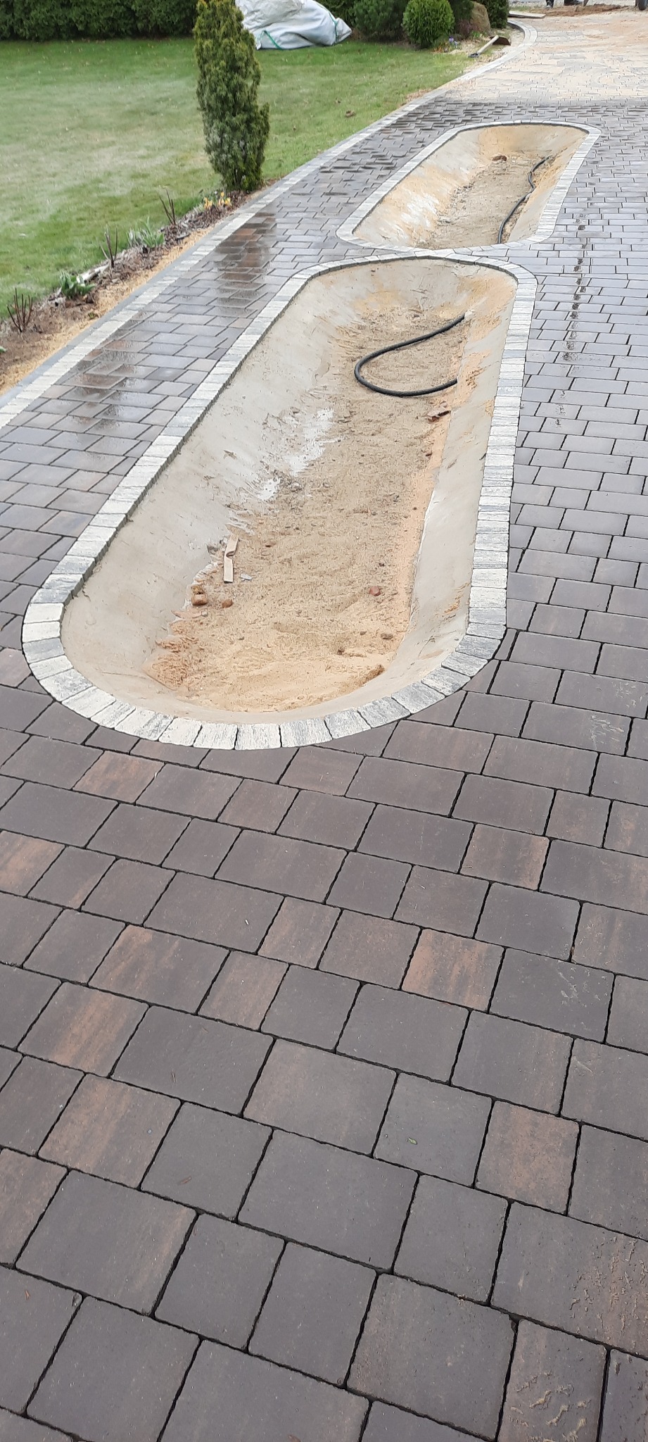 Ukończona ścieżka z kostki brukowej z dwoma wykopanymi, obramowanymi miejscami na rabaty kwiatowe, przygotowanymi do nasadzeń, z widocznym czarnym wężem ogrodowym w jednym z nich.