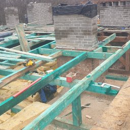 Antiza Usługi CIEŚIELSKO-DEKARSKIE - Konstrukcja dachu w trakcie budowy: widoczna drewniana więźba pomalowana na zielono, izolacja z wełny mineralnej, ceglany komin oraz narzędzia pracy dekarza, takie jak piła, poziomica i siekiera.