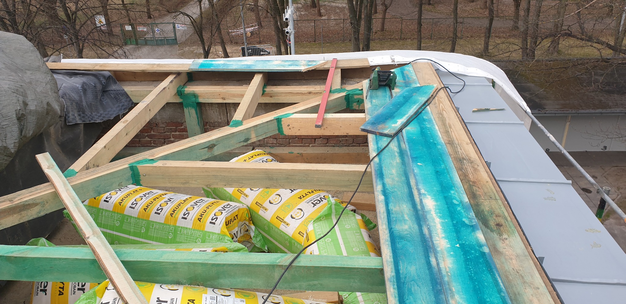 W trakcie budowy dachu: widoczna drewniana konstrukcja więźby dachowej, zabezpieczona zielonym impregnatem, częściowo pokryta folią, z izolacją termiczną w opakowaniach Isover Aku-Płyta, poziomica...