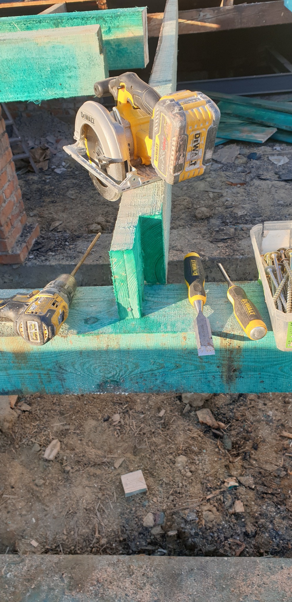 Piła tarczowa DeWalt na zielonej impregnowanej belce, obok wiertarka, dłuto i wkręty, prawdopodobnie podczas budowy więźby dachowej.