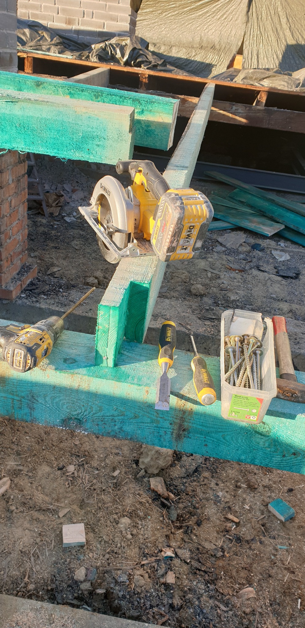 Fragment konstrukcji dachu w trakcie budowy, widoczne narzędzia DeWalt: piła tarczowa, wkrętarka, dłuto, śrubokręt oraz pojemnik ze śrubami do drewna.