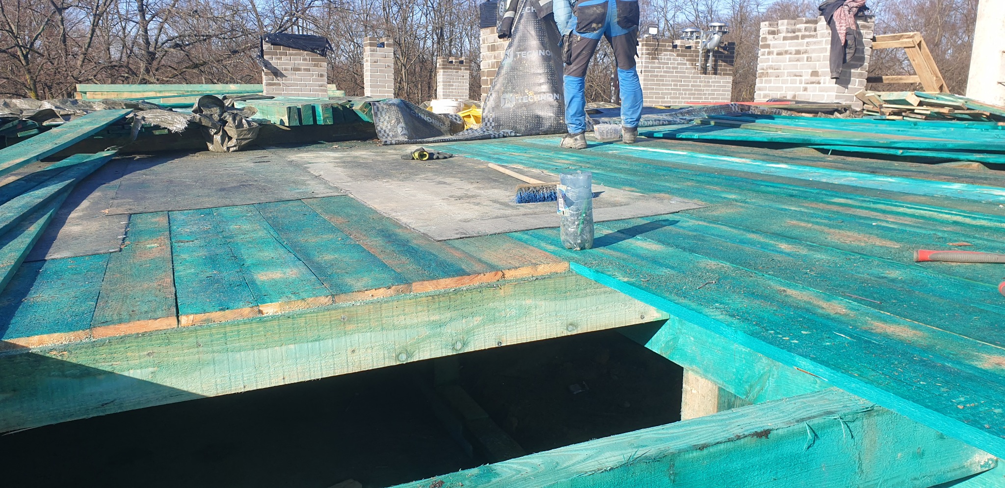 W trakcie montażu drewnianej konstrukcji dachu, widoczne impregnowane na zielono deski, fragment komina z cegły i pracownik w szelkach zabezpieczających.