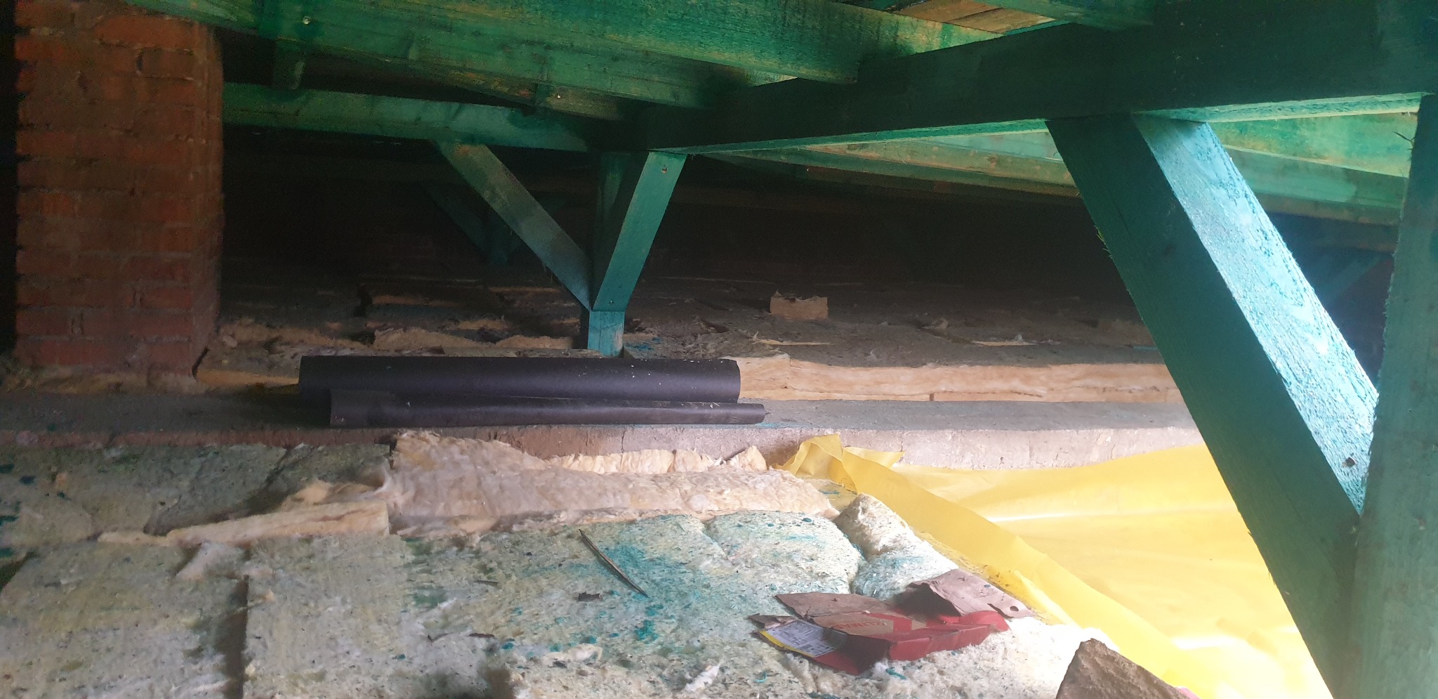 Wnętrze poddasza z widoczną konstrukcją więźby dachowej pomalowaną na zielono, fragmentem komina z czerwonej cegły, izolacją z wełny mineralnej i elementami folii budowlanej.