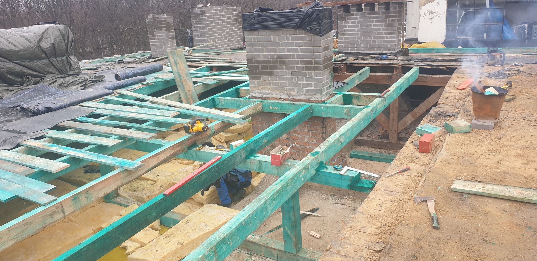 Konstrukcja dachu w trakcie budowy: widoczna drewniana więźba pomalowana na zielono, izolacja z wełny mineralnej, ceglany komin oraz narzędzia pracy dekarza, takie jak piła, poziomica i siekiera.